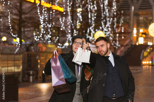 Two elegant young men standing in the street and holding the shopping bags and looking forward to the stores
