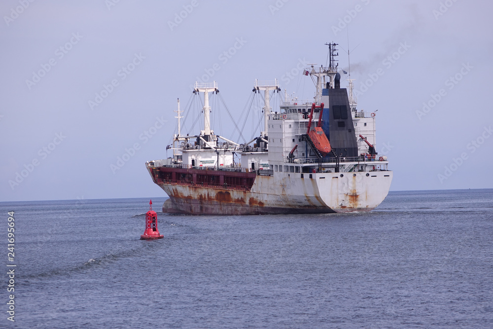 A rusty reefer ship leaving harbor next to a port buoy marker A rusty reefer ship leaving harbor next to a port buoy marker