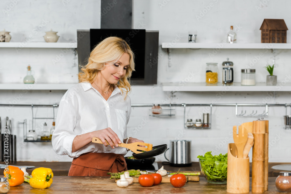 attractive middle aged woman cooking vegetables on frying pan in kitchen