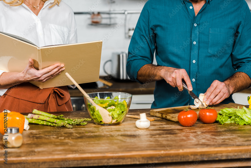 cropped image of mature wife and husband cooking together with recipe book in kitchen