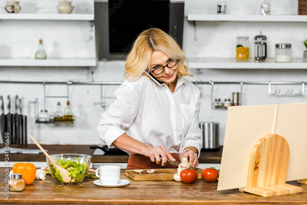 attractive mature woman talking by smartphone, cutting vegetables and looking at recipe book in kitchen attractive mature woman talking by smartphone, cutting vegetables and looking at recipe book in kitchen