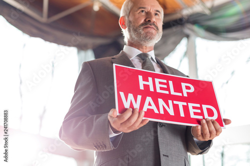 Low angle view of confident senior businessman holding help wanted sign while standing at office