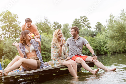 Cheerful shirtless man spraying water with squirt gun on friends sitting at pier over lake © moodboard