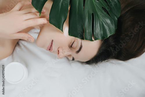 Portrait of a young long-haired girl applying cream on her face lying on the bed with a green leaf enjoying and relaxing . Lovely brunette with attractive appearance. Skin Care spa relax concept.