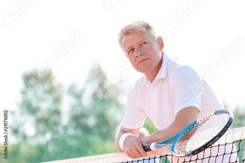 Thoughtful mature man holding tennis racket while leaning on net against clear sky