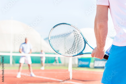 Midsection of mature man holding racket while playing with friends on tennis court