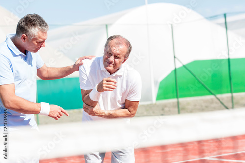 Canvas Print Mature man helping senior friend with elbow injury while playing tennis on sunny