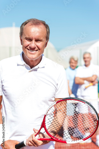 Portrait of smiling senior man holding tennis racket while standing against mature friends on court during sunny day © moodboard