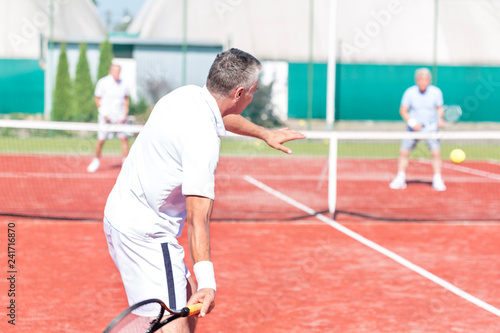 Man swinging racket while playing tennis doubles on red court during summer weekend