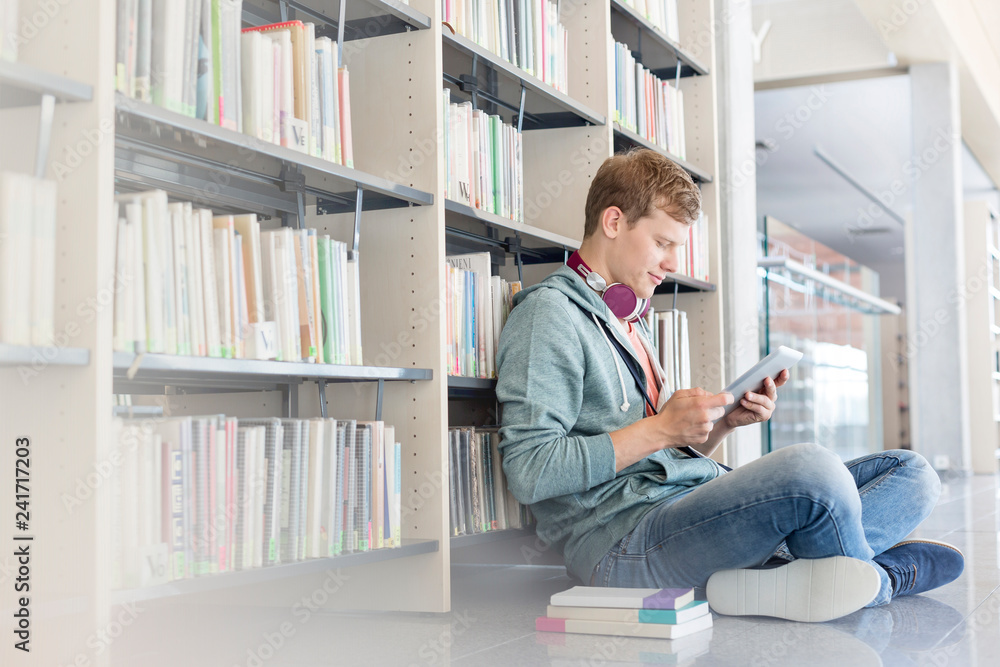 Student using digital tablet against bookshelf while sitting on floor at library - obrazy, fototapety, plakaty 