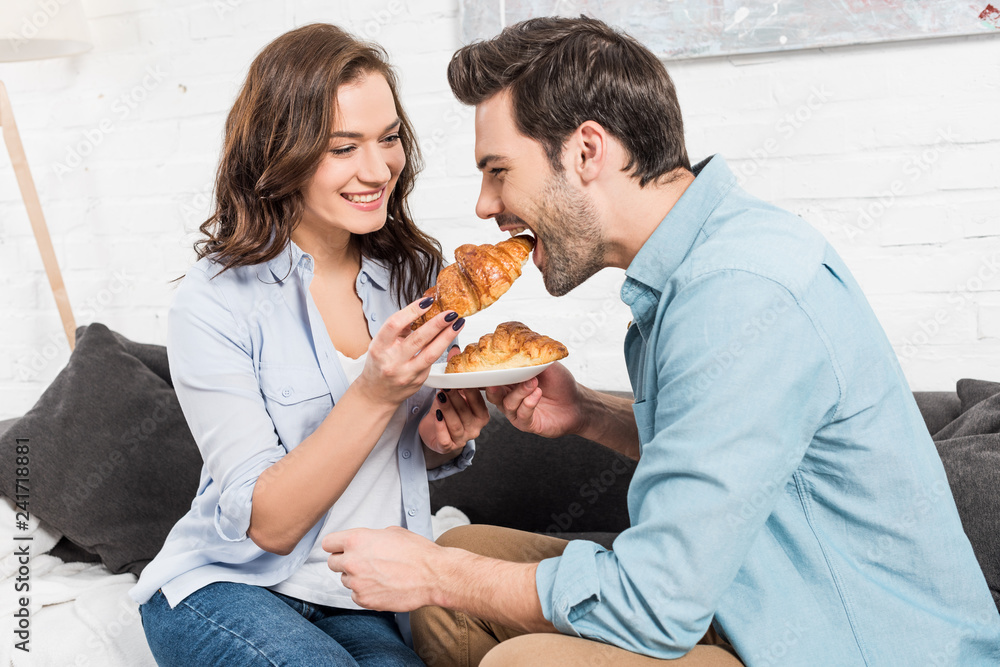 beautiful smiling woman feeding man with croissant during breakfast at home