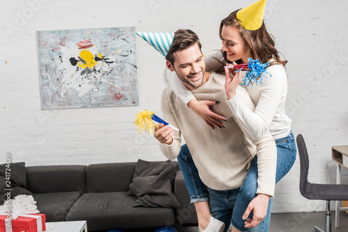 happy couple in party horns and party hats celebrating birthday while man giving piggyback ride to woman in living room