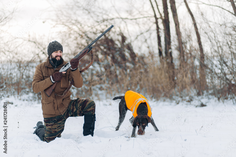 positive guy prepari ng a gun for shooting while his dog is looking something in the snow. full length photo.