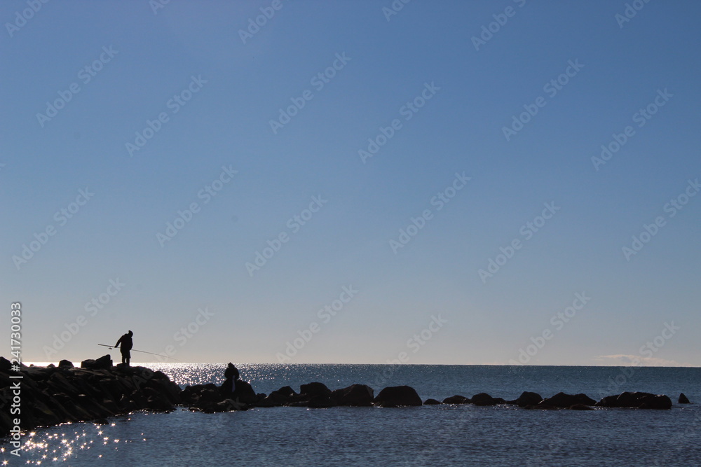 pescatori in controluce sugli scogli con cielo azzurro e raggi del sole riflettono sull'acqua