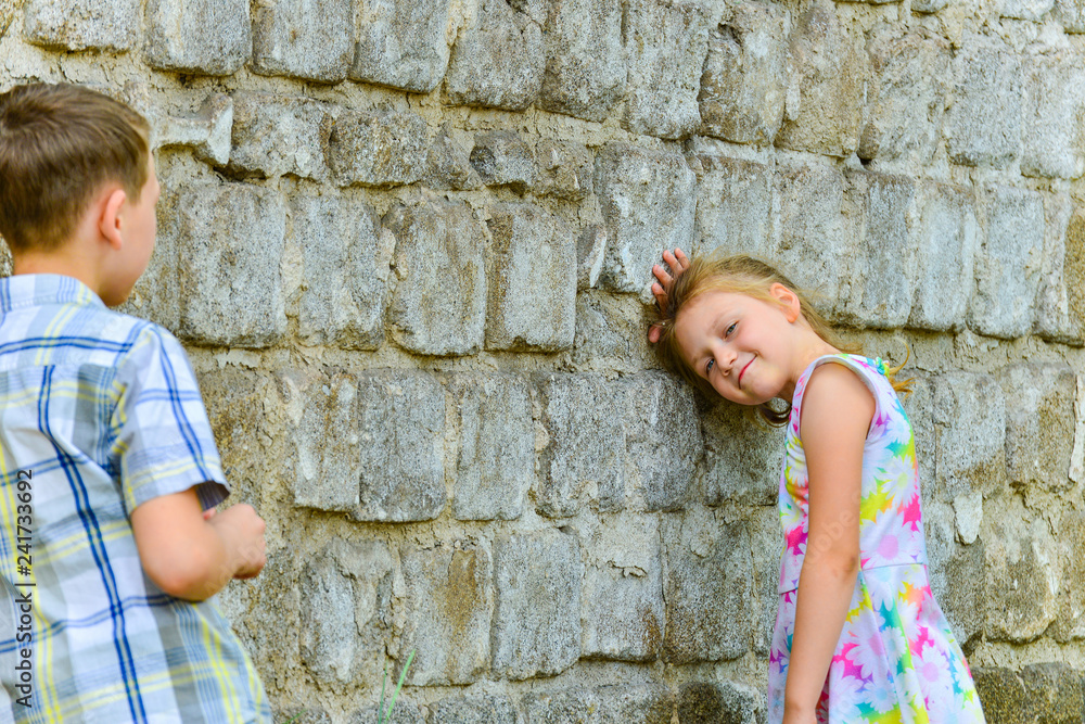 The boy is leaning against the brick wall, and the girl is posing for the camera on the street. The boy is leaning against the brick wall, and the girl is posing for the camera on the street.