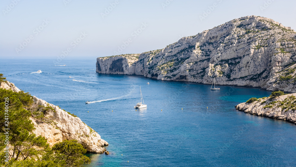 Panoramic view of the cap Morgiou on the mediterranean shore near Marseille, France, with motorboats cruising and sailboats mooring in the blue waters of the calanque de Morgiou on a sunny day.