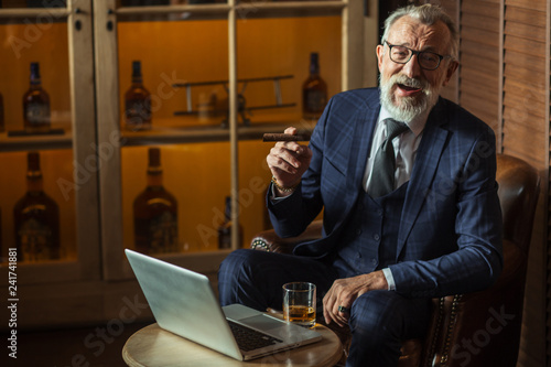 Bearded old-aged playwriter looking at camera with a cheerful smile while writing his new novel using laptop, while sitting in restaurant. He smoking a cigar and sipping whiskey photo