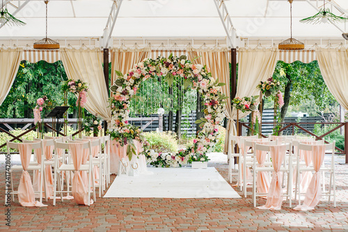 Beautiful wedding set up. Area of the wedding ceremony. Round arch, white chairs decorated with flowers, greenery. Cute, trendy rustic decor. Part of the festive decor, floral arrangement.