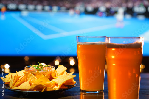 2 glasses of beer with nachos and salsa on a dark wooden table. Sports bar, tennis on a background, high resolution