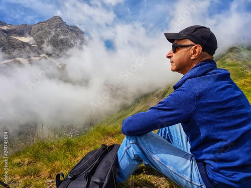 A man sits on a hillside and admires the tops of mountains in the clouds.