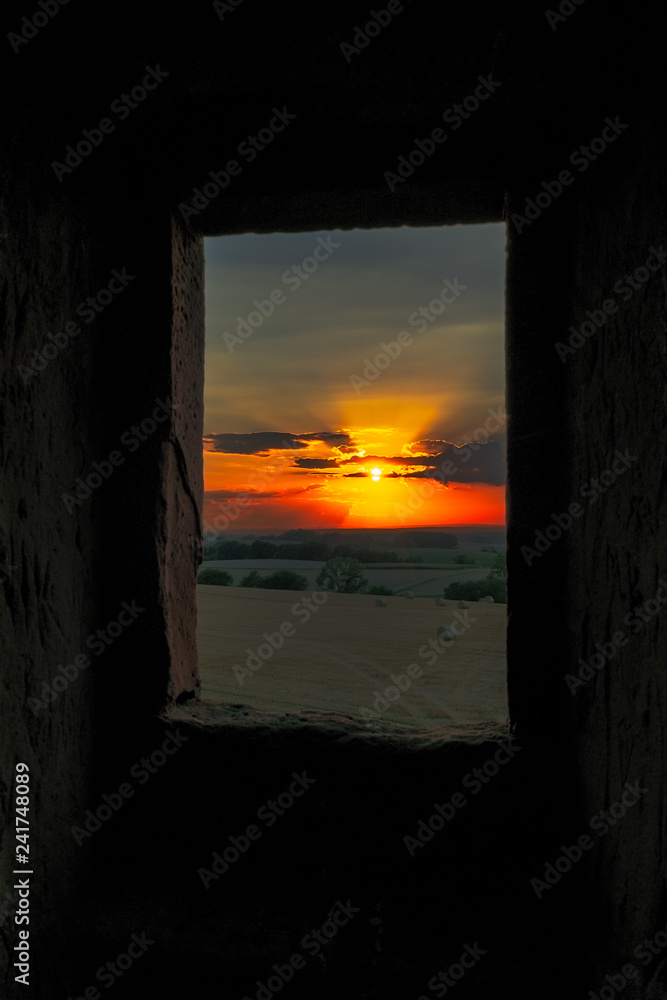 Sonnenuntergang durch ein Burgfenster Fotografiert.Im Vordergrund Feld und Wiesen die Sonne steht in der Mitte vom Bild.