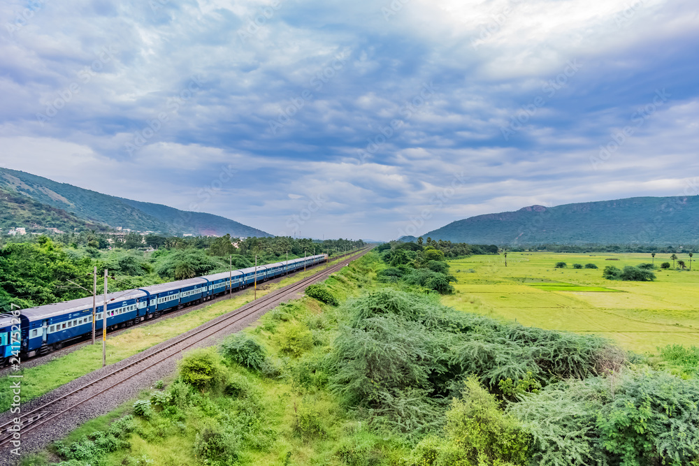 awesome view of indian railway running on track goes to horizon in green landscape under blue sky with clouds.
