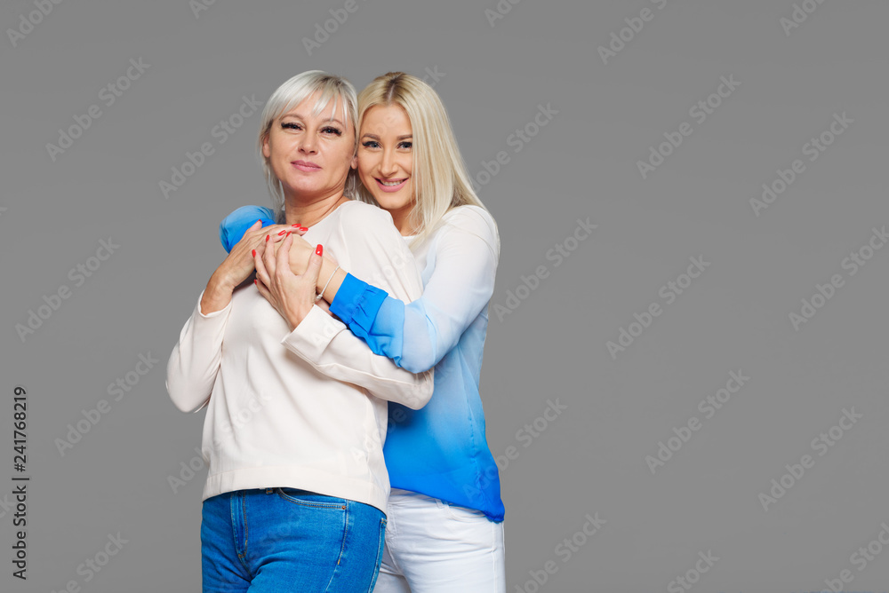 Teen blond daughter and mature mother on grey background hugging Teen blond daughter and mature mother on grey background hugging