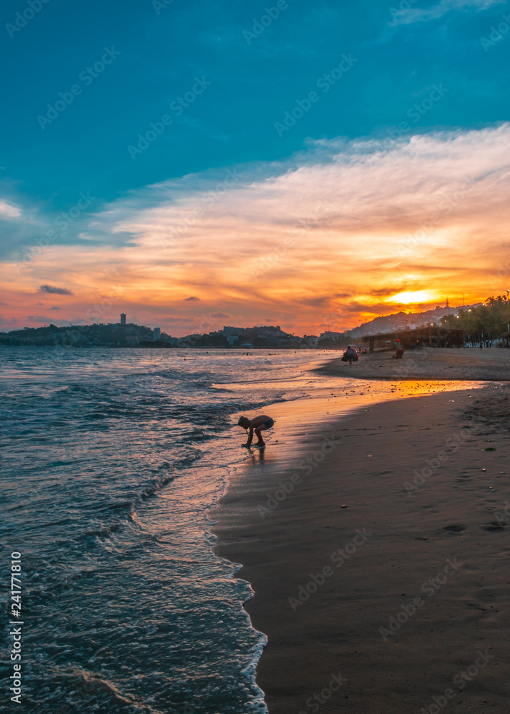 Longshot of a beautiful orange sunset and a kid playing at the edge of the sea in the beach of Acapulco.