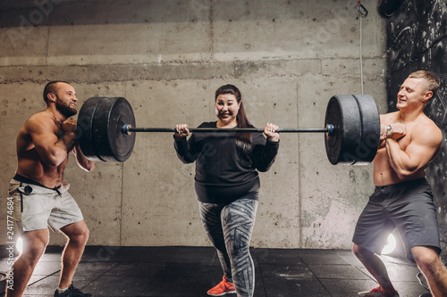 Fat crazy sweaty woman workout with men at gym. guys help plump girl to lift the weight. bodybuilding