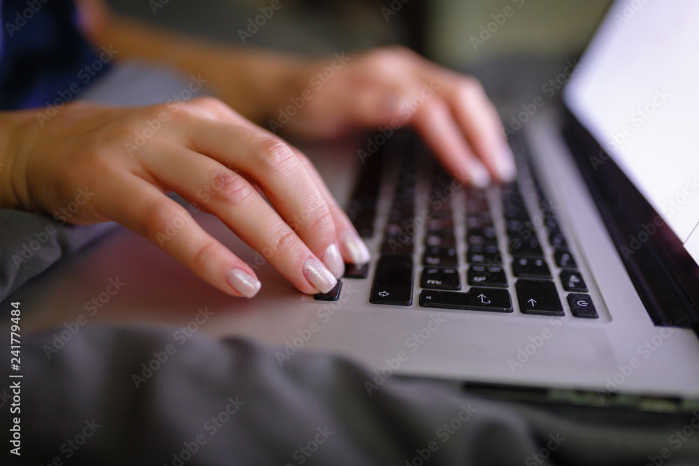 Close-up of woman's hand using laptop with blank screen on bed in home interior. The light from the screen illuminates the female hands on the laptop keyboard. Freelancer works at home in the evening