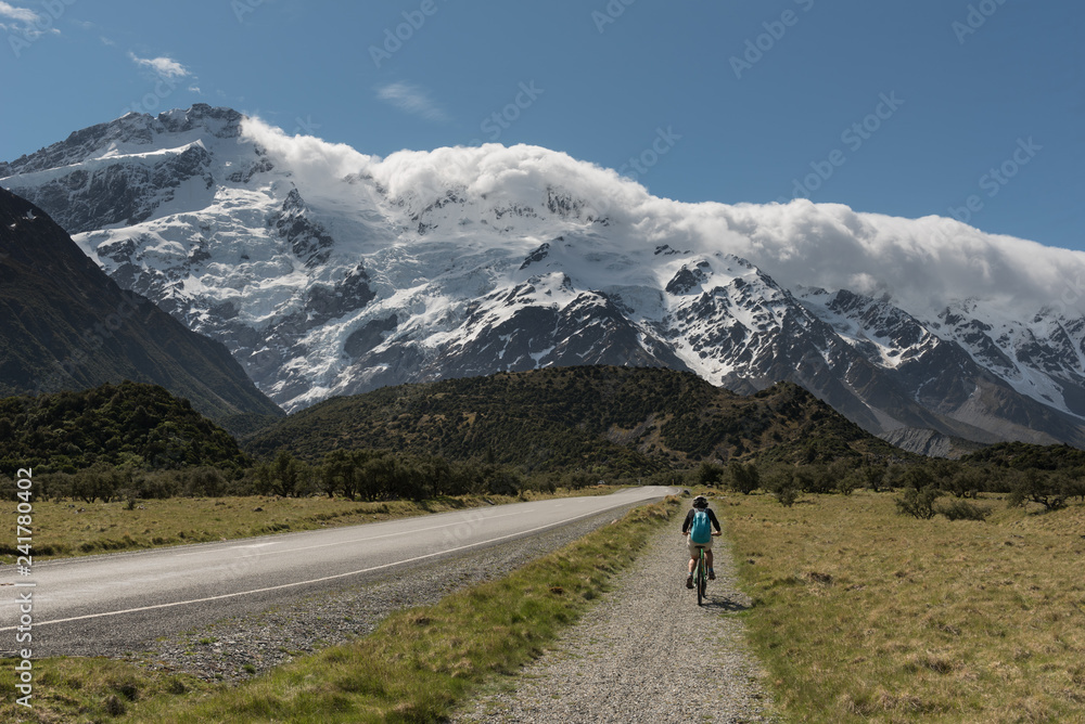 Female, baby boomer cyclist cycling on a bike path towards the snow capped and glacier covered Southern Alps, Aoraki/Mount Cook National Park, New Zealand.