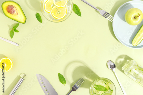 A glass beaker, a bottle of cucumber water, fruits and cutlery on a light green background. Minimalistic creative concept. Copy space. Top view.