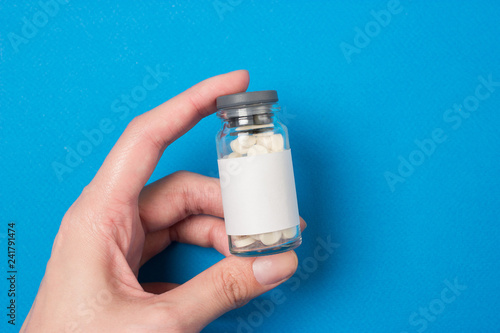 A glass flask with white tablets inside and a rubber cap that is held in a female hand. Paper blue background. Top view.