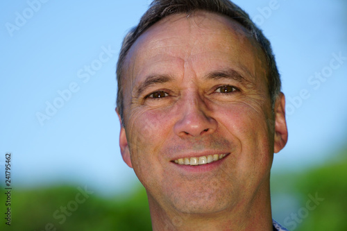 An outdoor portrait of a caucasian man wearing a blue shirt and smiling at the camera.