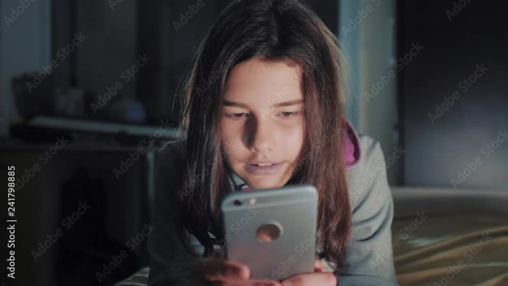 Teenage girl online shopping looks on her smartphone in bed during the night. little teen hood girl writes a message chatting in social media messenger night evening indoors smartphone on lifestyle Teenage girl online shopping looks on her smartphone in bed during the night. little teen hood girl writes a message chatting in social media messenger night evening indoors smartphone on lifestyle