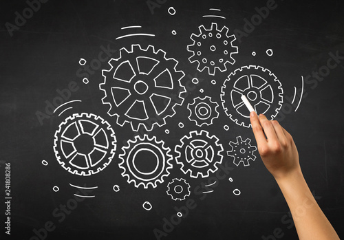 Female hand holding white chalk in front of a blackboard with gears drawn on it