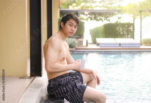 asian man sitting on the edge of a swimming pool and holding cup of coffee © Nattapol