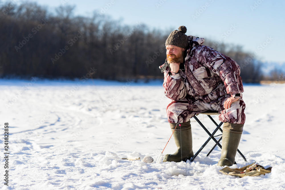 Bearded european man is boring while he fishing from ice hole on the river at winter day time
