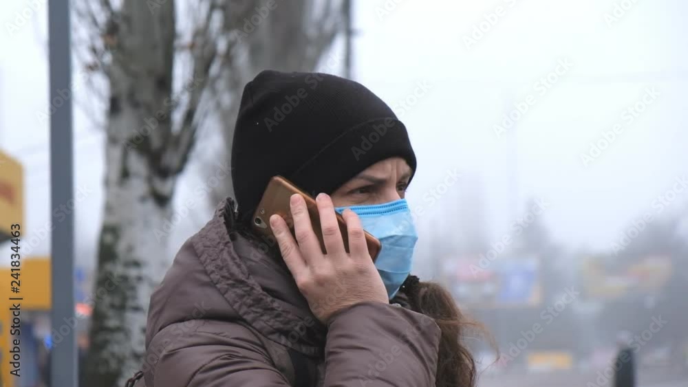 a young woman is standing on a city street in dense smog in a medical mask and talking on the phone.