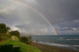 Big double rainbow over sea and coastline, green plants on bank, person walk on beach, cloudy sky