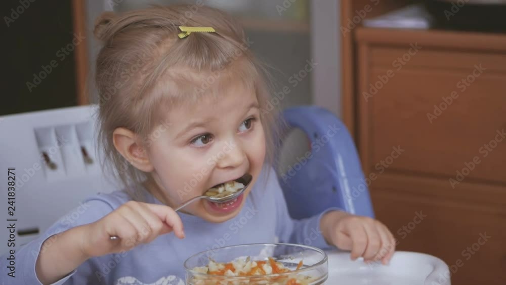 A little girl eats a spoonful of vegetable soup soiling her face and carefully watches TV A little girl eats a spoonful of vegetable soup soiling her face and carefully watches TV