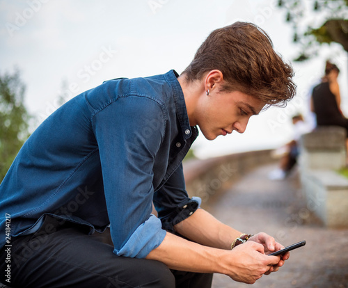 Handsome trendy man wearing shirt sitting and typing on cell phone  outdoor in city setting in day shot