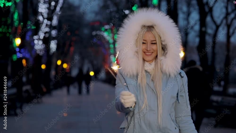 Cheerful young woman in a warm fluffy jacket is standing and holding a sparkler in her hand outdoor, Christmas lights background. Girl is fun at a party with a firework in her hands, city landscape. Cheerful young woman in a warm fluffy jacket is standing and holding a sparkler in her hand outdoor, Christmas lights background. Girl is fun at a party with a firework in her hands, city landscape.
