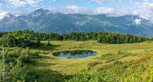 View of a mountain glade with a transparent mirror lake, and tourists, surrounded by grass and trees and high mountains with snow-capped peaks in the distance. Caucasus, Russia
