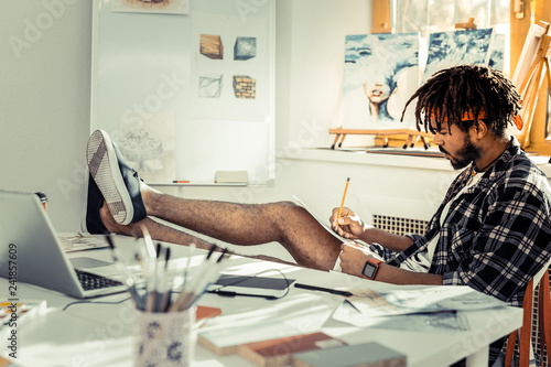 Bearded artist with dreadlocks putting his legs on table while drawing