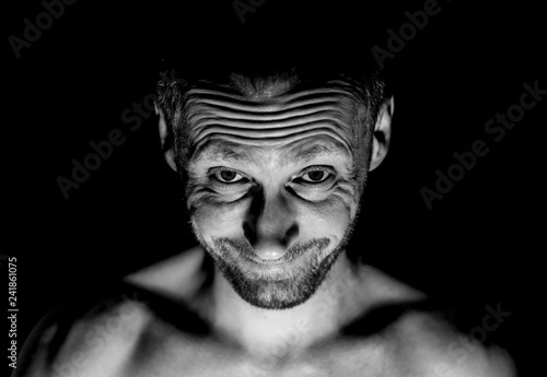 Portrait of unshaven adult caucasian man. He smiles like maniac and seems like madness. Black and white shot, low-key lighting. Isolated on black.