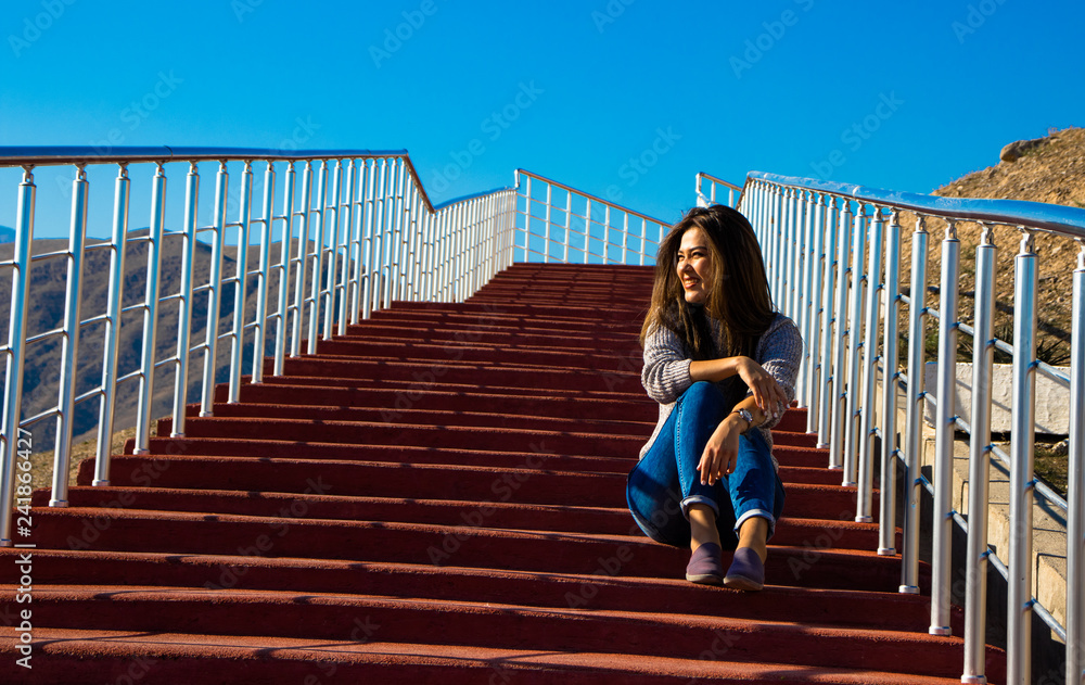 a young woman sitting on red stairs and smiling at midday on a background of mountains and blue sky a young woman sitting on red stairs and smiling at midday on a background of mountains and blue sky