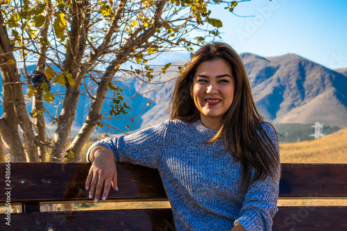 a young woman sitting on a bench and smiling on a background a tree covered with yellow leaves and a beautiful view on Kopet Dag mountains in Turkmenistan  