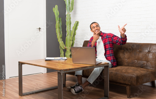 African american man with laptop in the living room pointing with the index finger and looking up