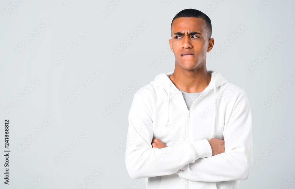 Dark-skinned young man with white sweatshirt having doubts and with confuse face expression while bites lip. Questioning an idea on grey background Dark-skinned young man with white sweatshirt having doubts and with confuse face expression while bites lip. Questioning an idea on grey background